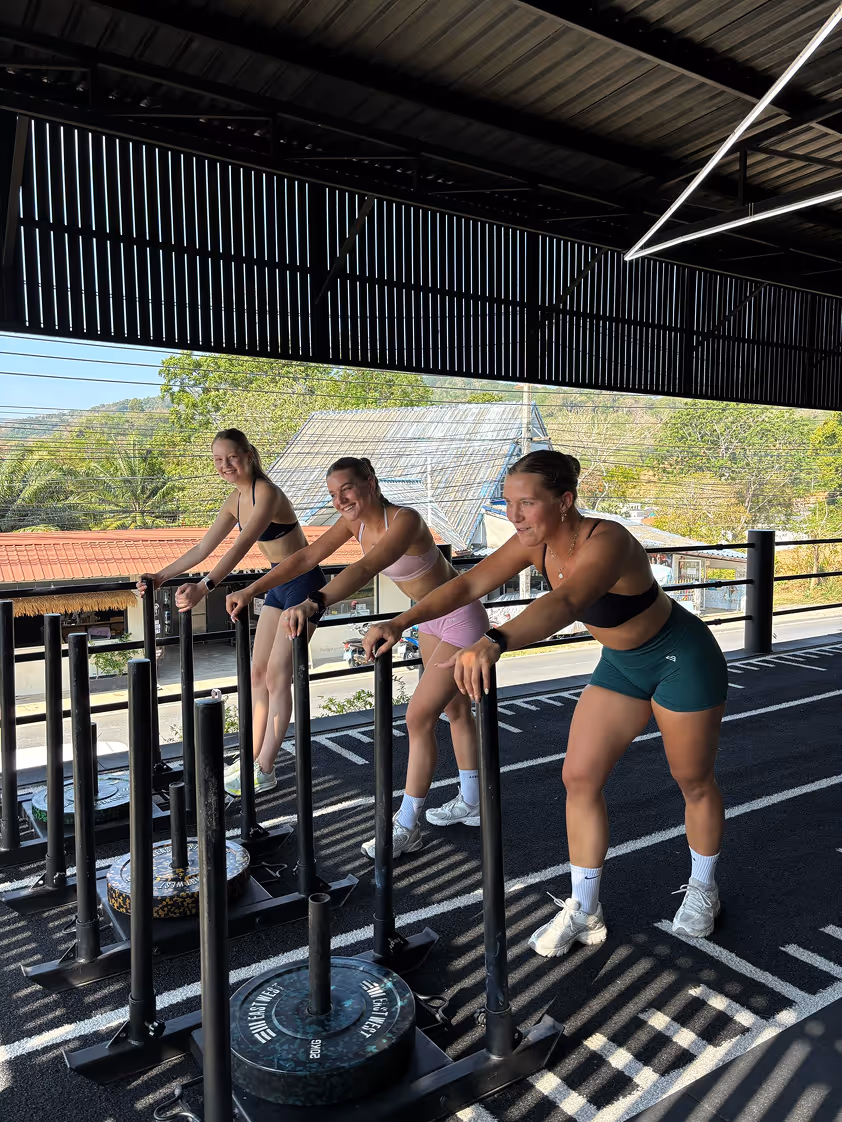 Drie vrouwen in sportkleding leunen op trapeze-sledes in een open fitnessruimte met uitzicht op bomen en huizen.