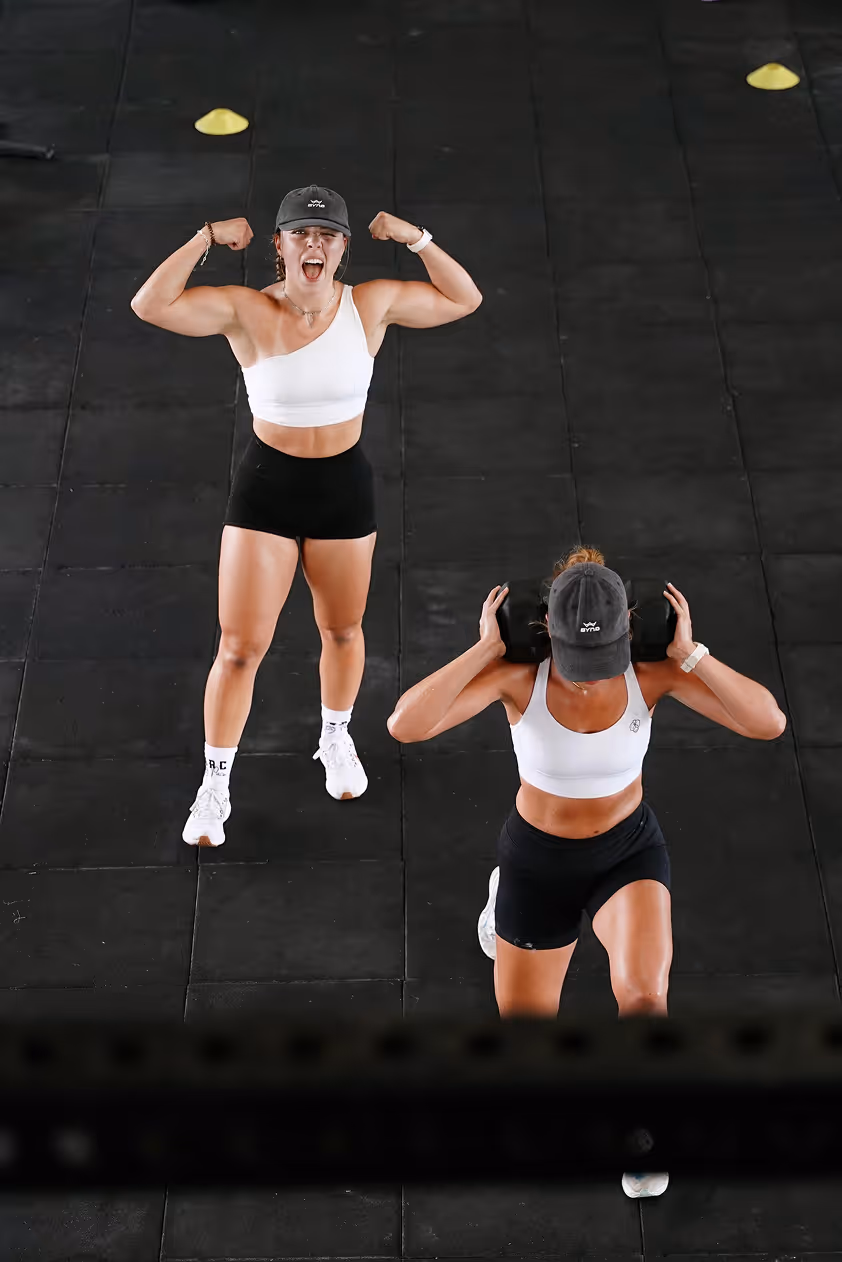 Zes vrouwen in bikini poseren vrolijk op een zandstrand met turquoise water en rotsachtige eilanden op de achtergrond.