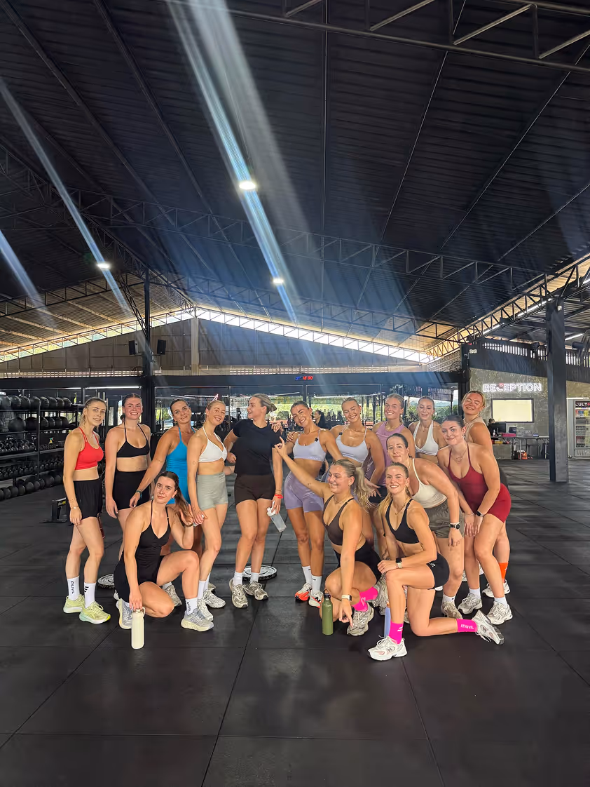 Groep vrouwen in sportkleding poseert in een sportschool met hoog plafond en rekken met gewichten.