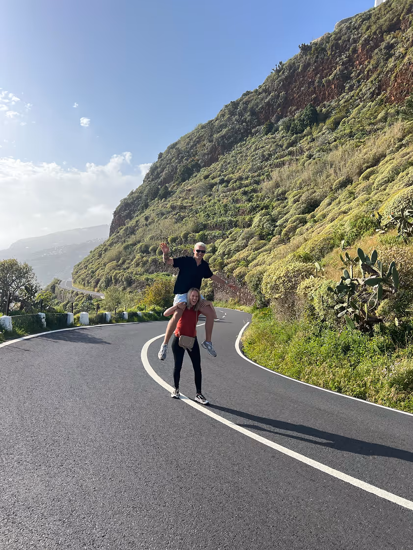 Twee mensen lachen op een bochtige bergweg, een vrouw draagt een man op haar schouders, omgeven door groene heuvels onder een blauwe lucht.