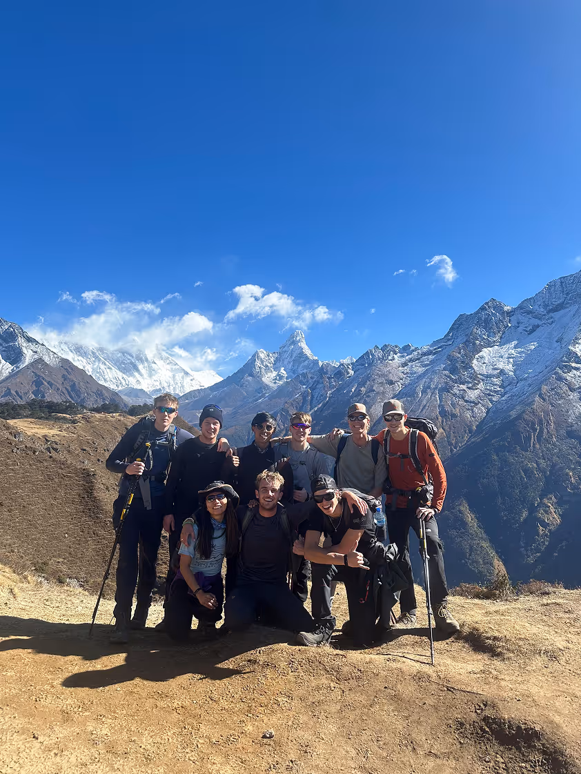 Een groep van negen wandelaars poseert op een bergpad met besneeuwde bergen en een heldere blauwe lucht op de achtergrond.