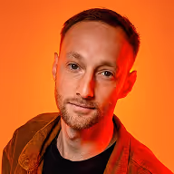 Portret van een man met kort bruin haar en baard, met een oranje achtergrond.
