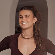 Vrouw met lang gekruld haar lacht en draagt meerdere kettingen en een donker topje met een diepe halslijn.