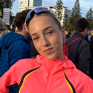Jonge vrouw met lichtroze jas en zonnebril op haar hoofd poseert buiten bij een evenement.