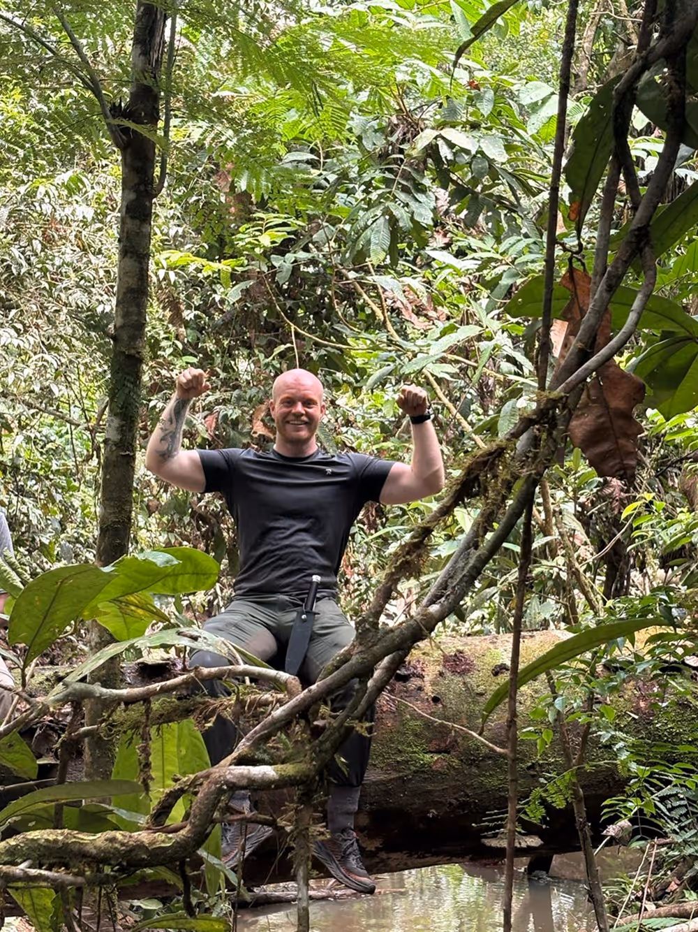 Man in zwart shirt en kaki broek zit op een omgevallen boomstam in een dichtbegroeid tropisch bos met opgeheven armen en een glimlach.