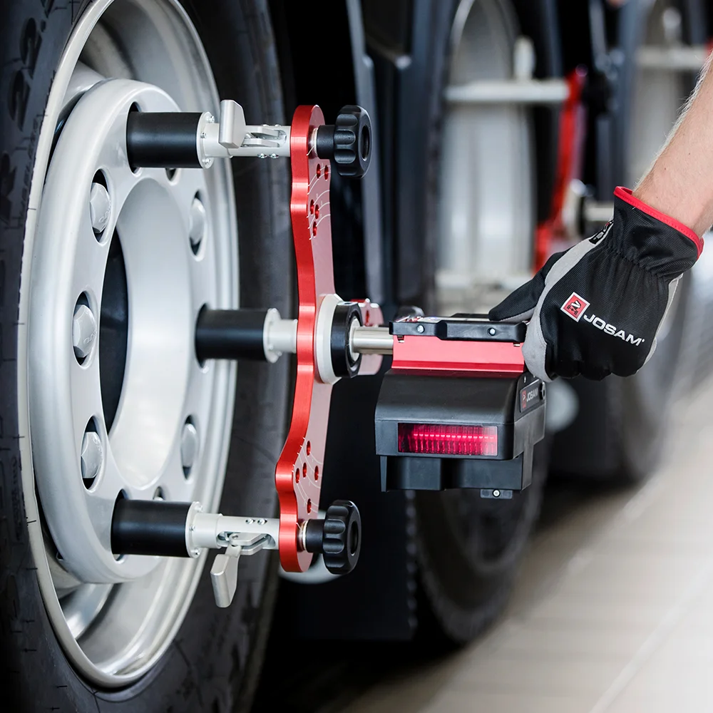 Close-up of a hand wearing a Josam glove adjusting a red and black wheel alignment tool mounted on a truck tire.
