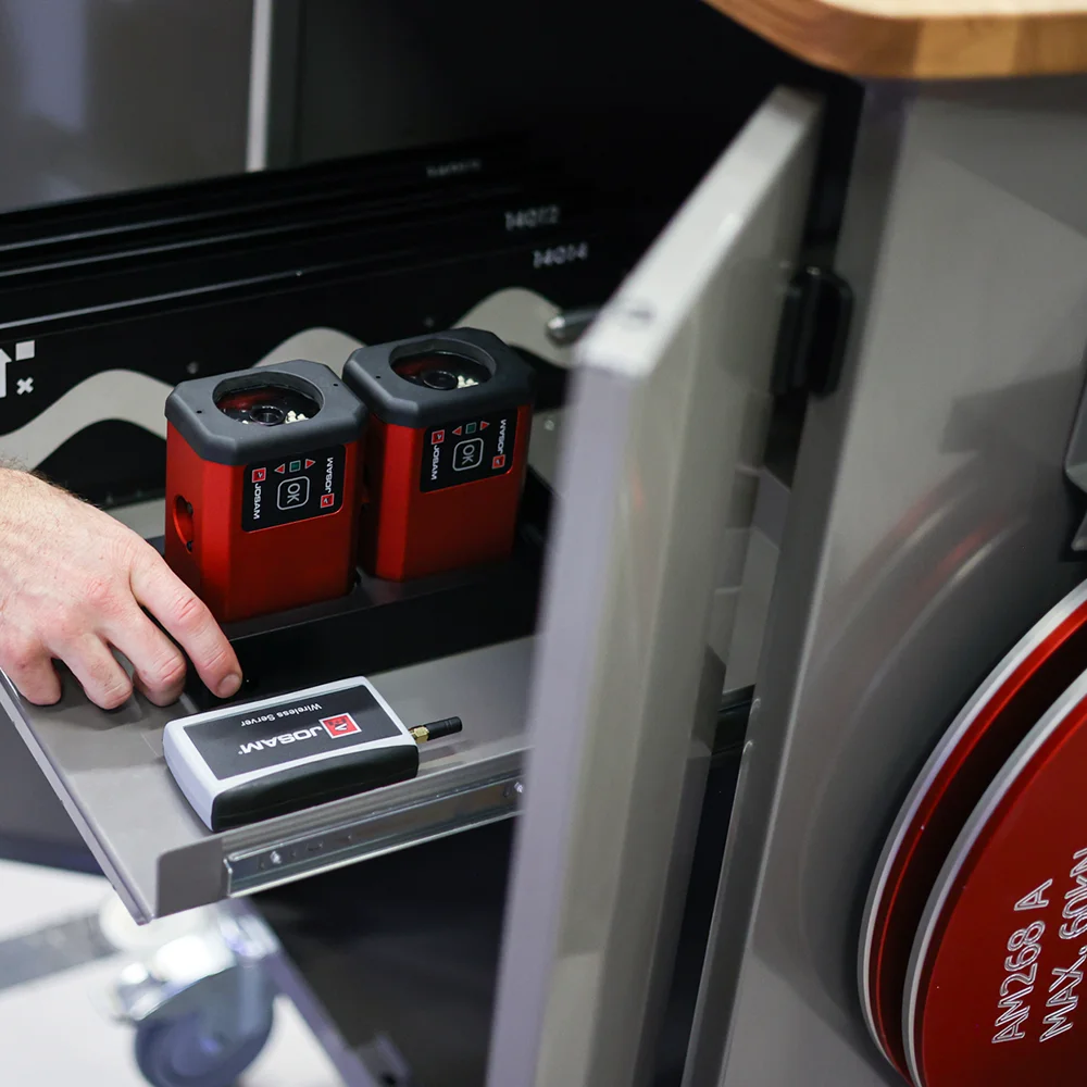 Hand reaching for two red JOSAM devices and a wireless service unit on a metal tray inside a gray equipment cabinet.