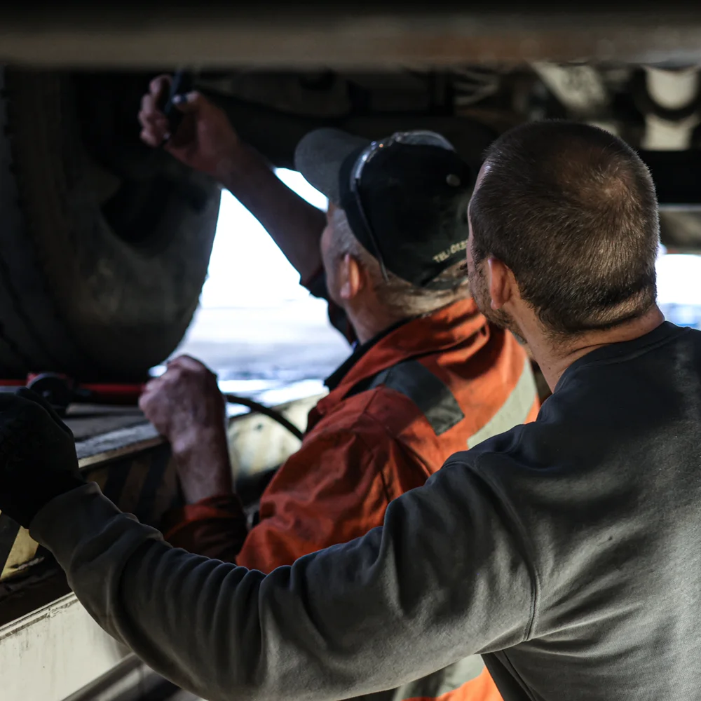 Two men inspecting the underside of a vehicle, one holding a flashlight while the other observes closely.
