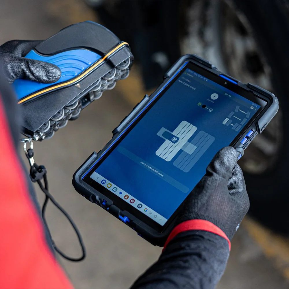 Person wearing black gloves holding a rugged tablet displaying a tire alignment app and a blue handheld tire tread depth gauge.