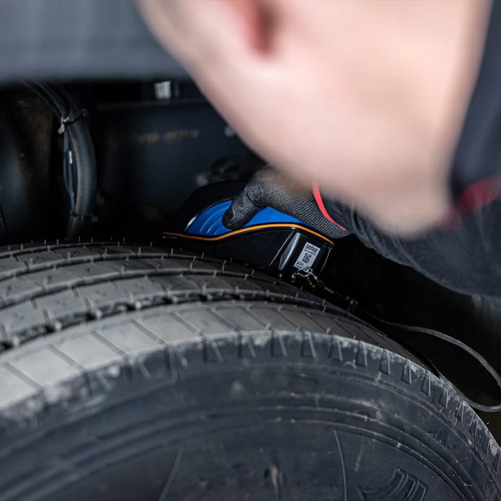 Person wearing a black glove holding a blue and black device near the tire in a vehicle's wheel well.