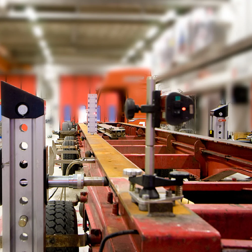 Close-up of a precision measuring setup on a red metal chassis frame in a workshop with an orange truck in the background.
