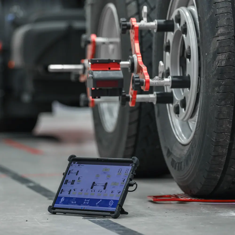 Vehicle wheel alignment sensors attached to truck wheels with a tablet displaying alignment data on the floor.