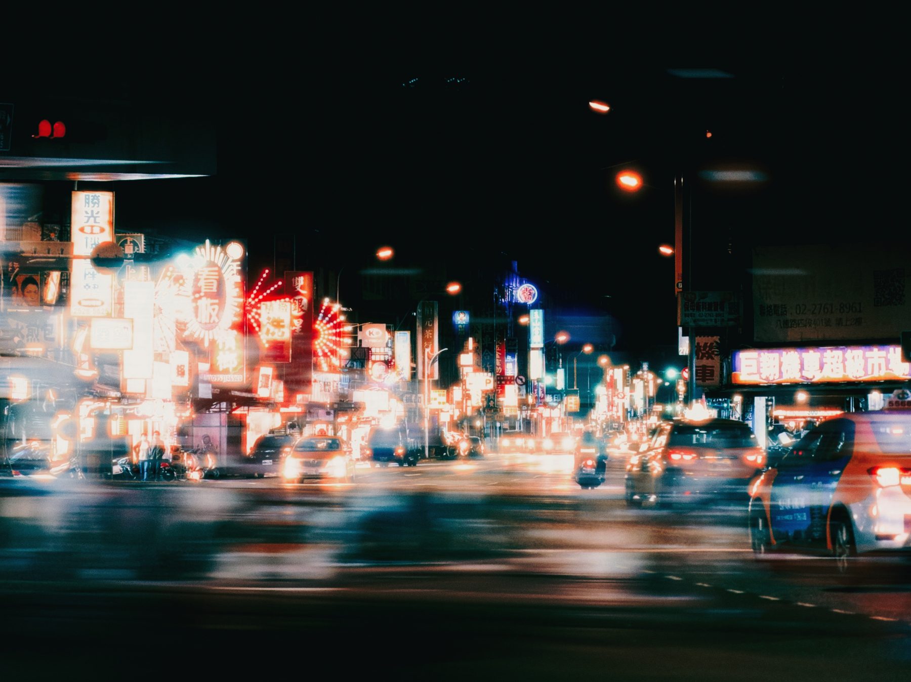 A night city street where car and shop lights are stretched into long neon streaks