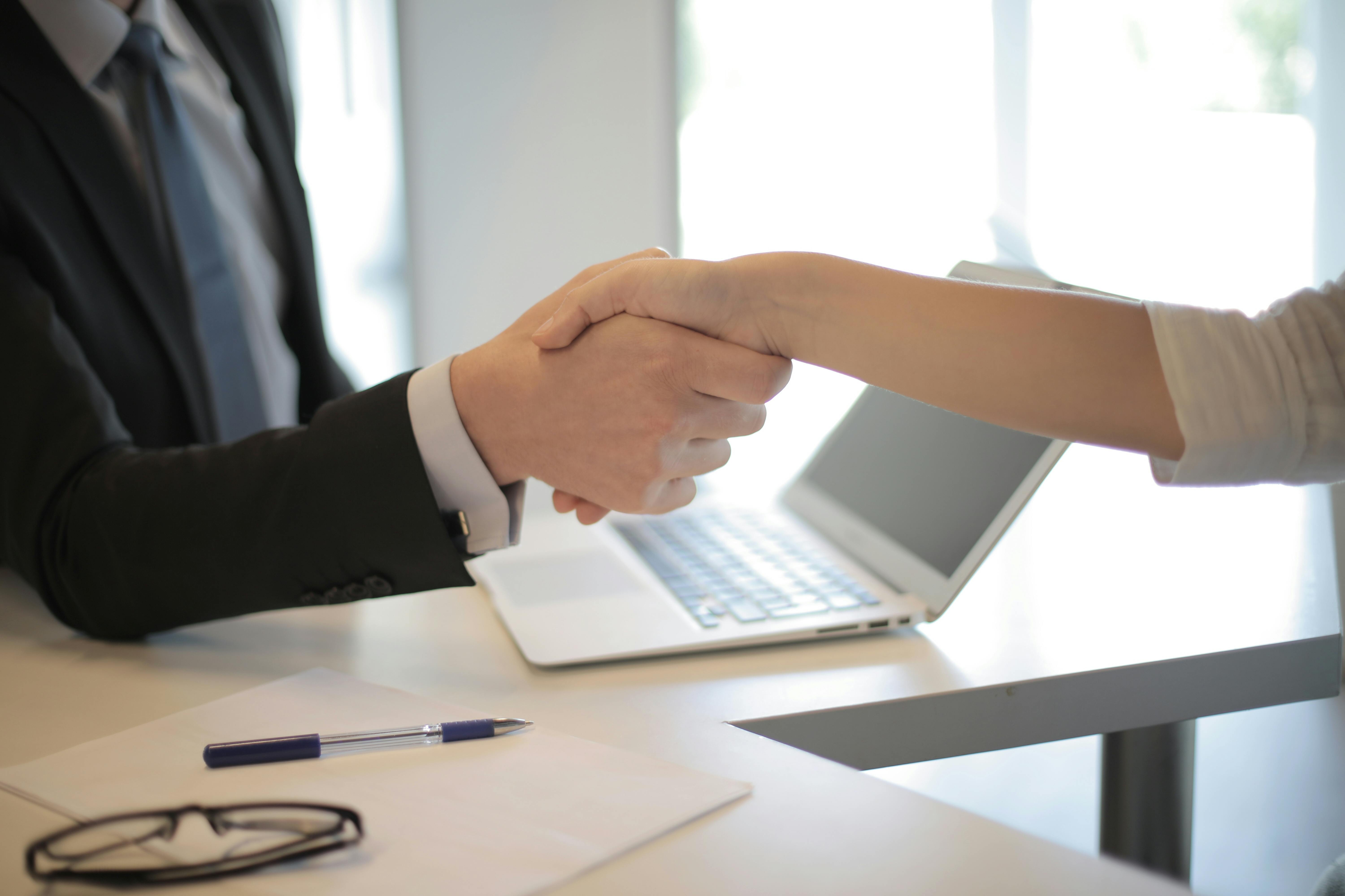 Business buyer and seller shaking hands across a desk after signing financial agreement, modern office background, professional setting, confident expressions.