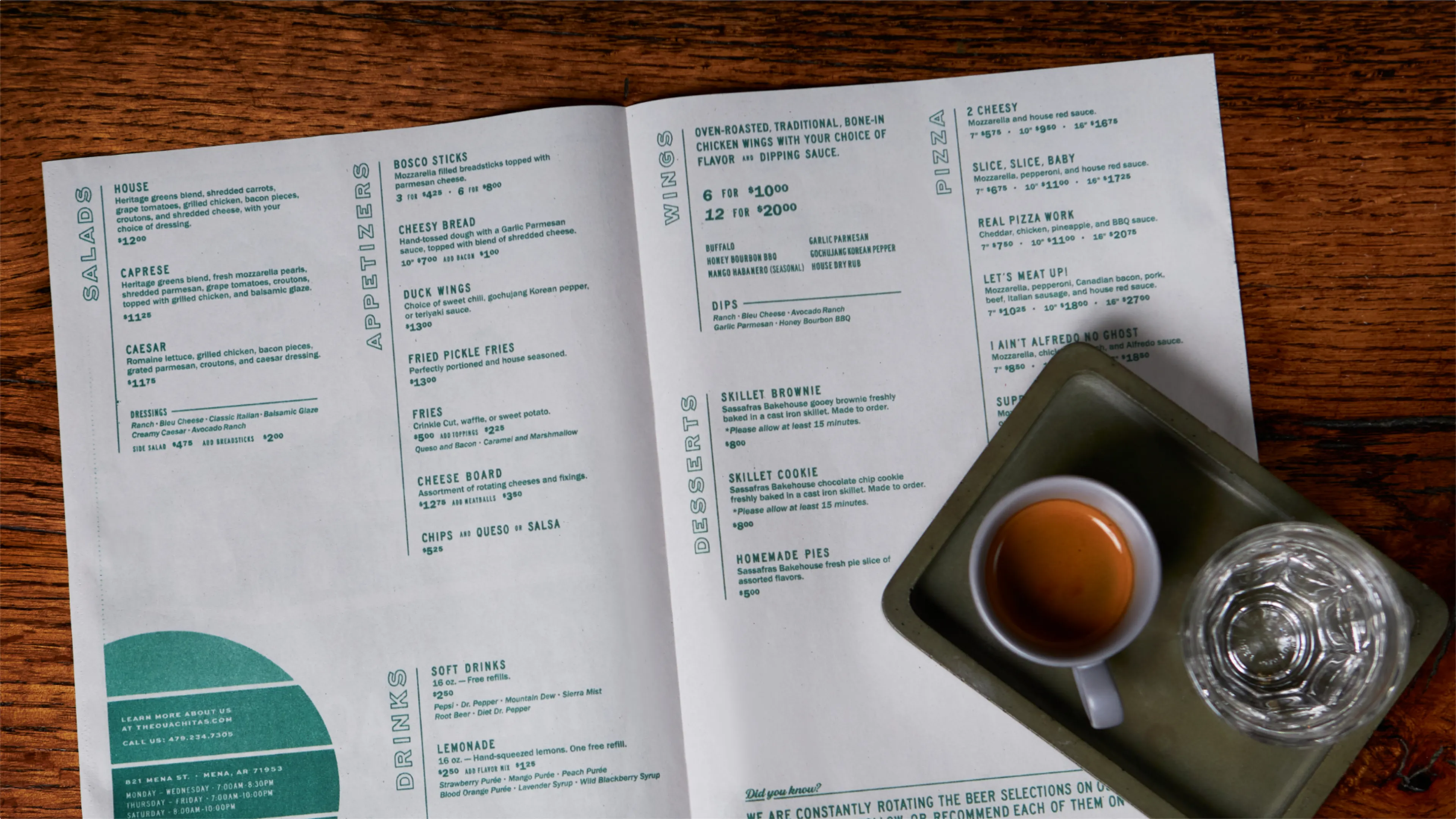The Ouachitas restaurant menu open spread showing salads, appetizers, wings, pizza, and desserts alongside espresso and water glass on rustic wood table, designed by Unbound Collective