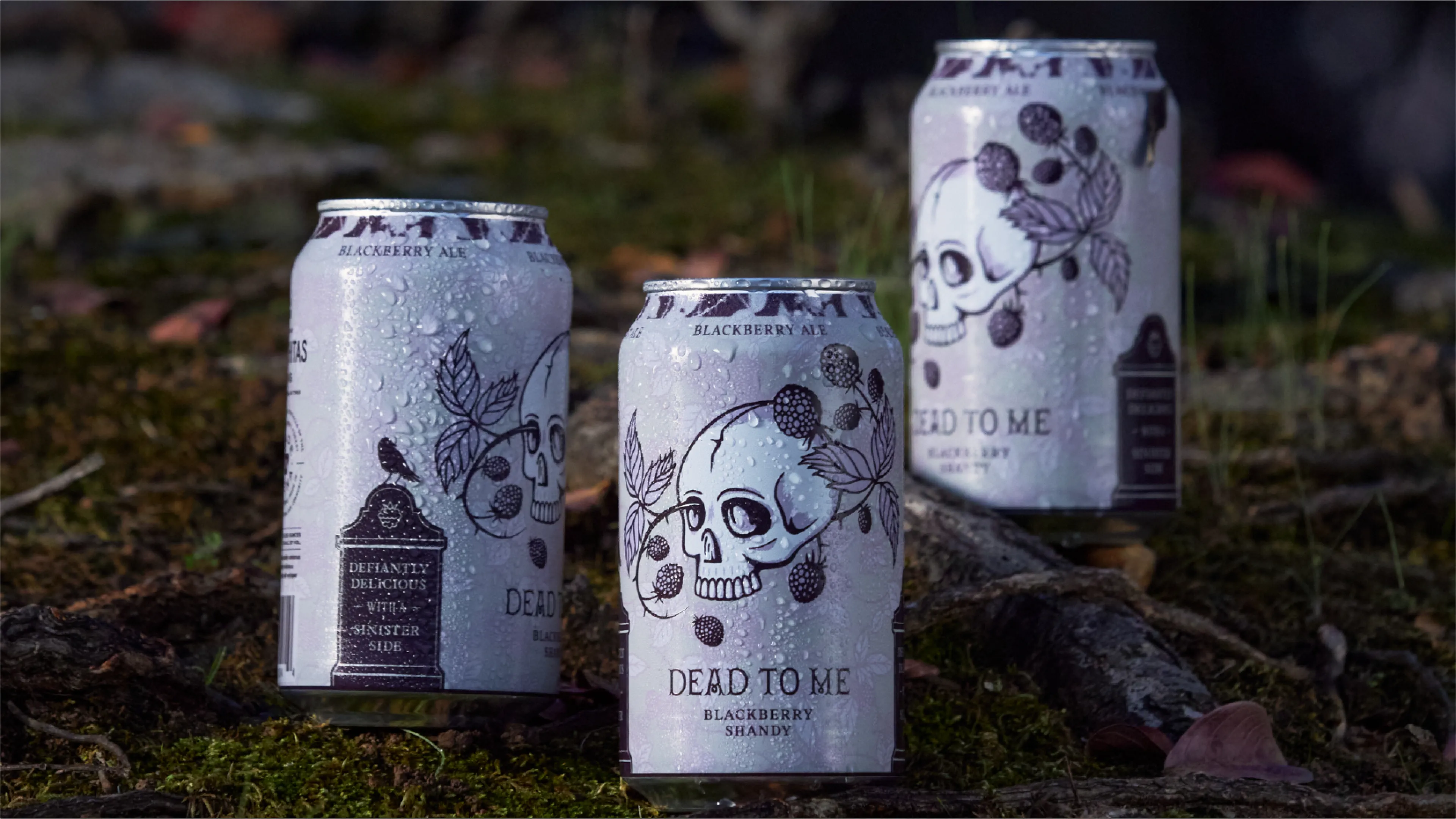 The Ouachitas Dead to Me blackberry shandy beer cans with skull and blackberry illustration resting on mossy rocks outdoors, can design by Unbound Collective