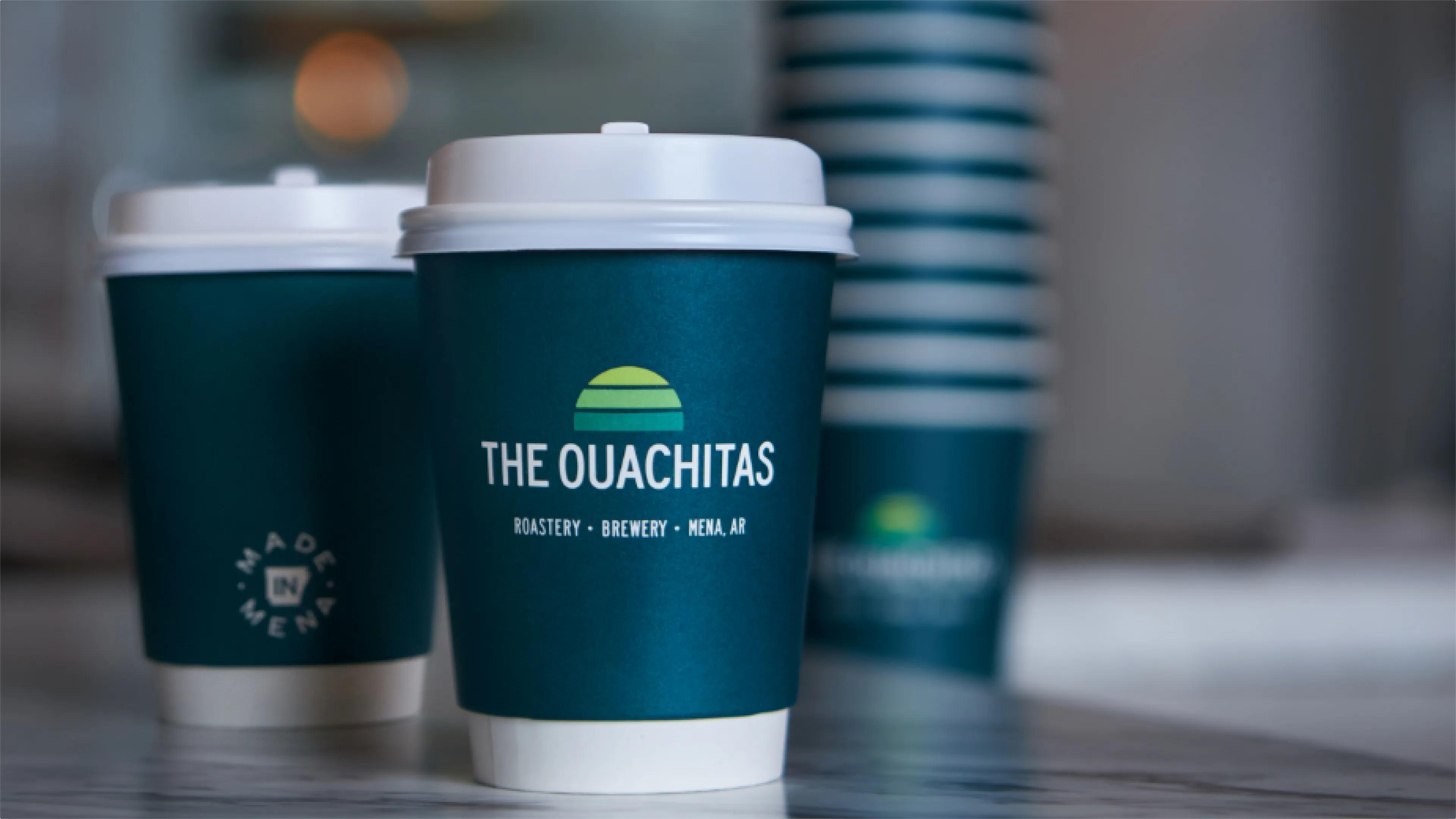 The Ouachitas teal branded to-go coffee cups with sunrise logo reading Roastery Brewery Mena AR and Made in Mena stamp, packaging designed by Unbound Collective