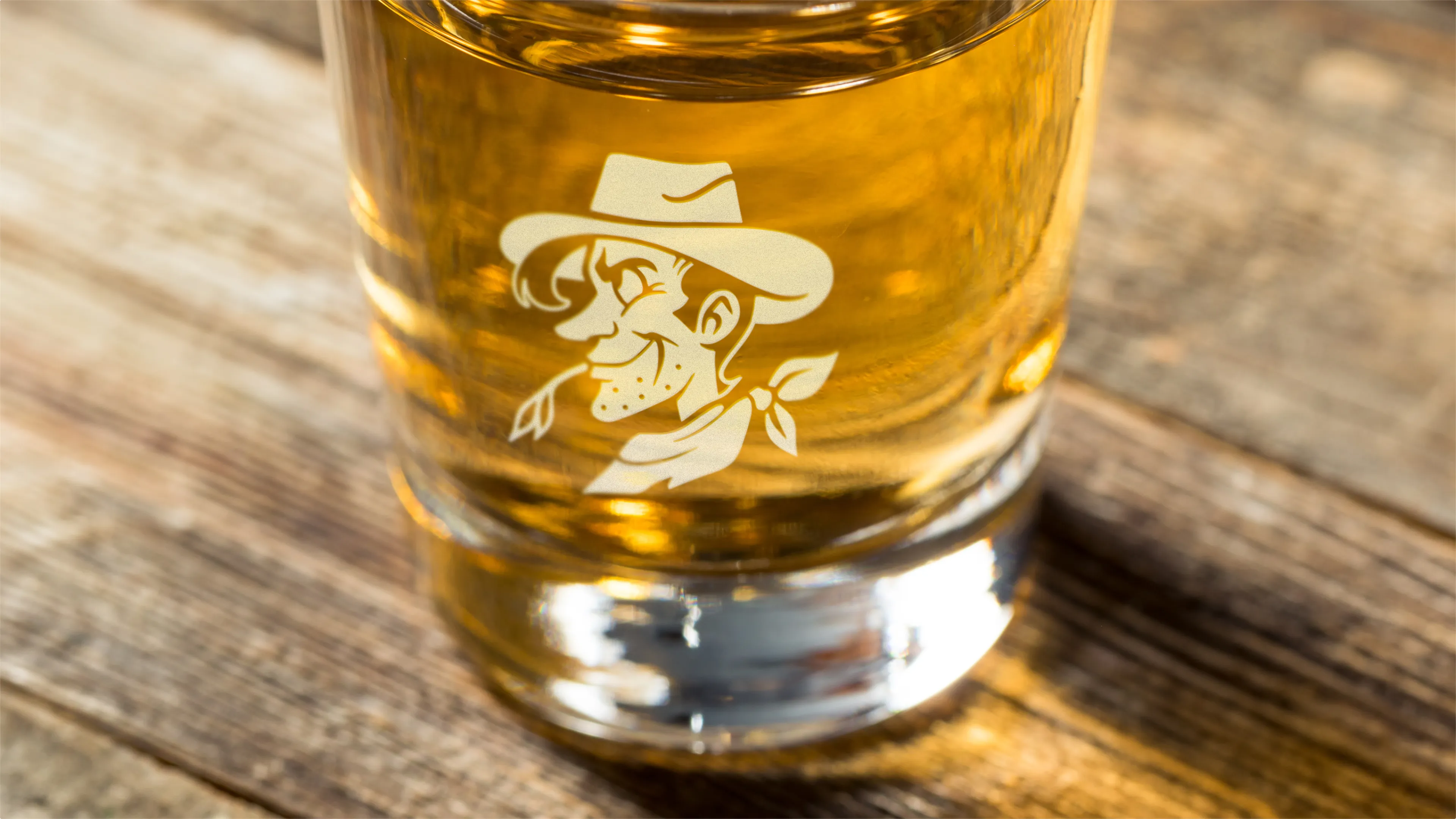 Poco's Beer Wine and Spirits etched pint glass with vintage cowboy mascot logo filled with golden beer on wood surface, merchandise designed by Unbound Collective