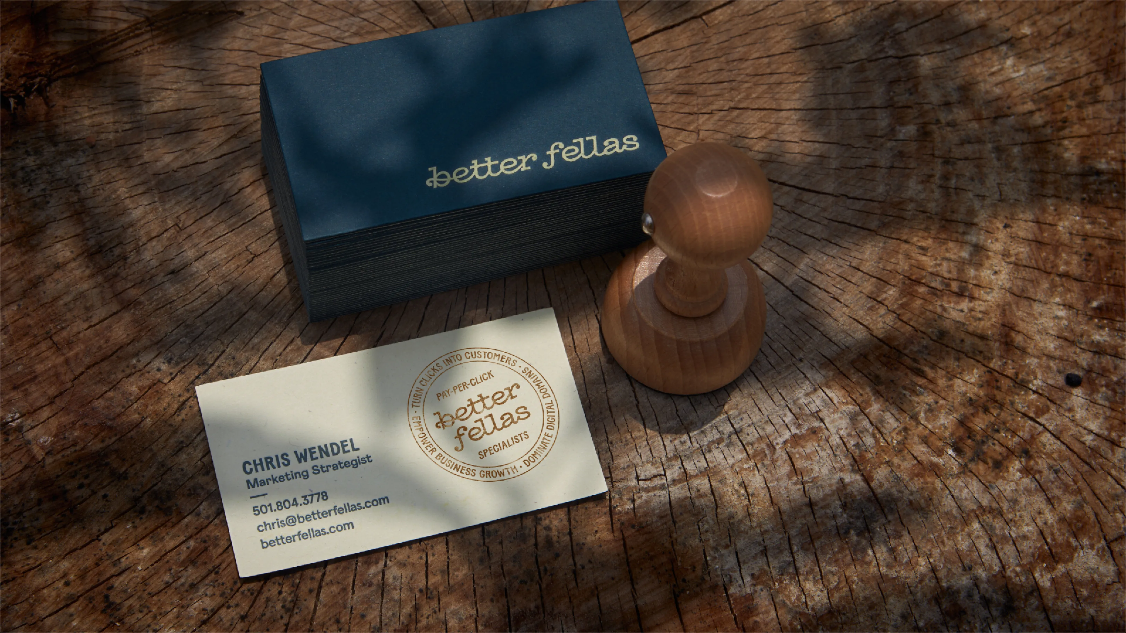 Better Fellas navy and cream business card with PPC specialist circular stamp alongside wooden rubber stamp on rustic wood stump surface, brand identity designed by Unbound Collective