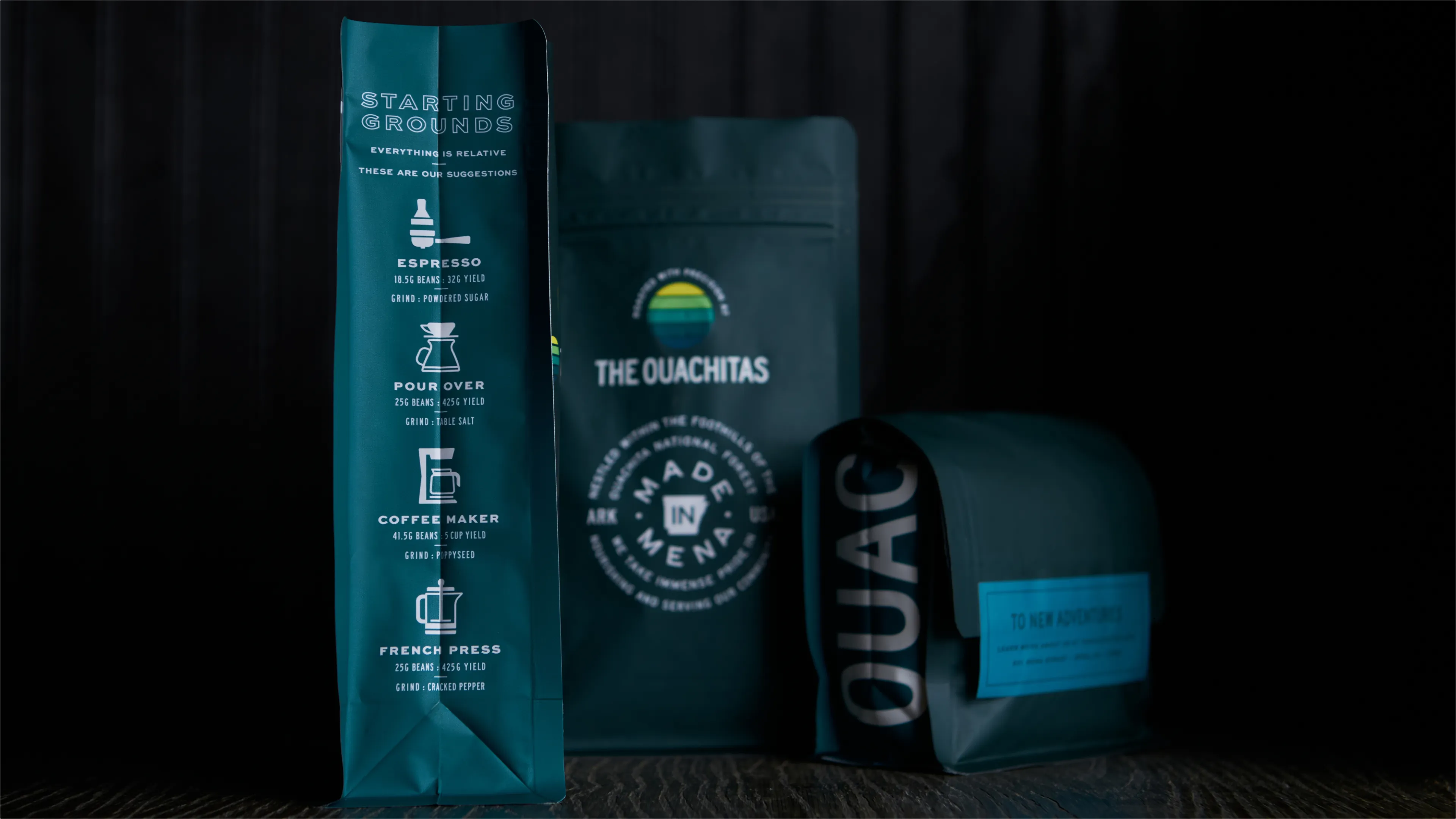 The Ouachitas Starting Grounds teal coffee brewing guide bag alongside Made in Mena stamp bag and teal Ouachitas branded bag on dark surface, packaging designed by Unbound Collective
