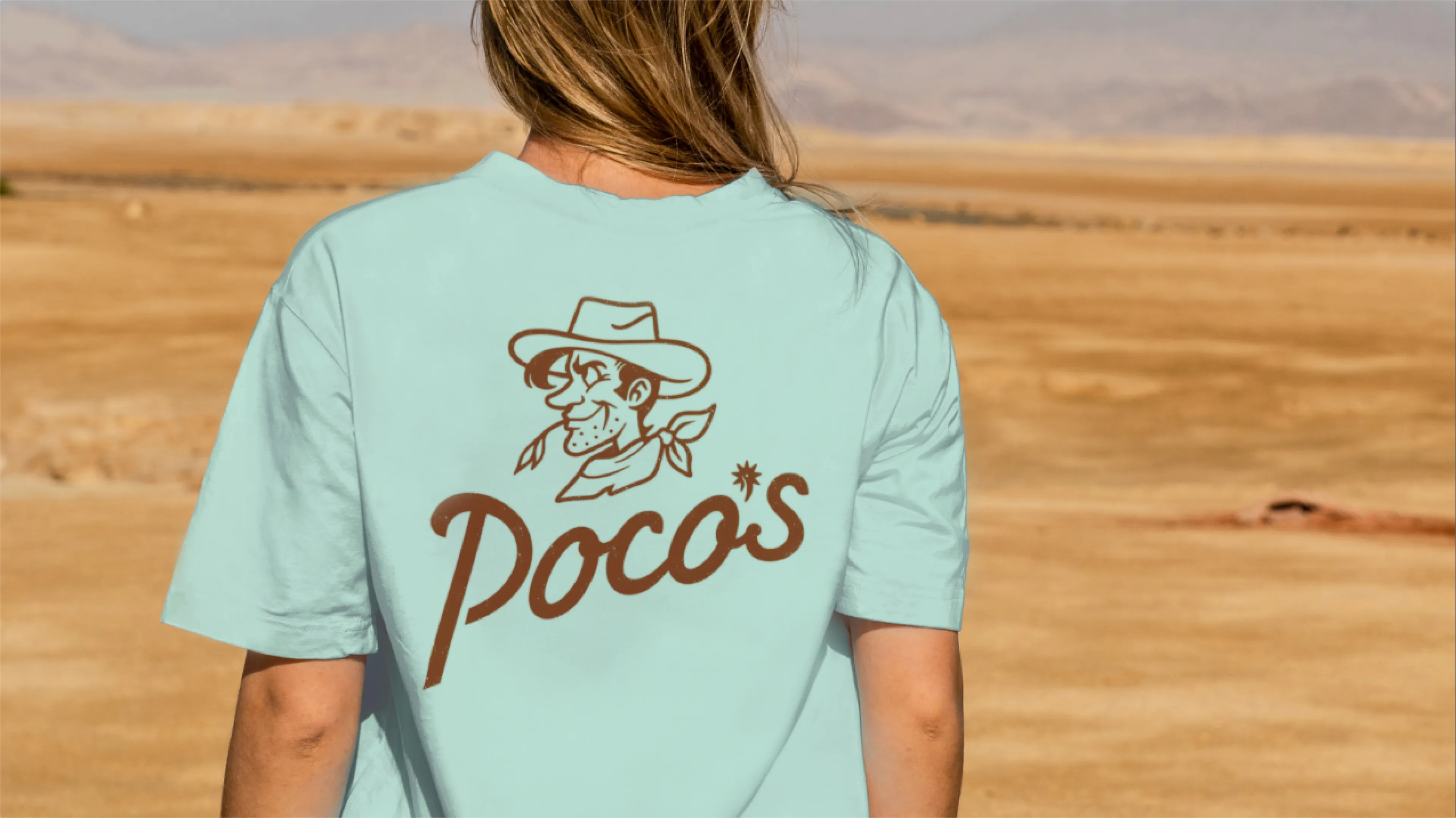 Poco's Beer Wine and Spirits vintage cowboy mascot logo on light blue t-shirt worn outdoors with desert landscape background, merch design by Unbound Collective