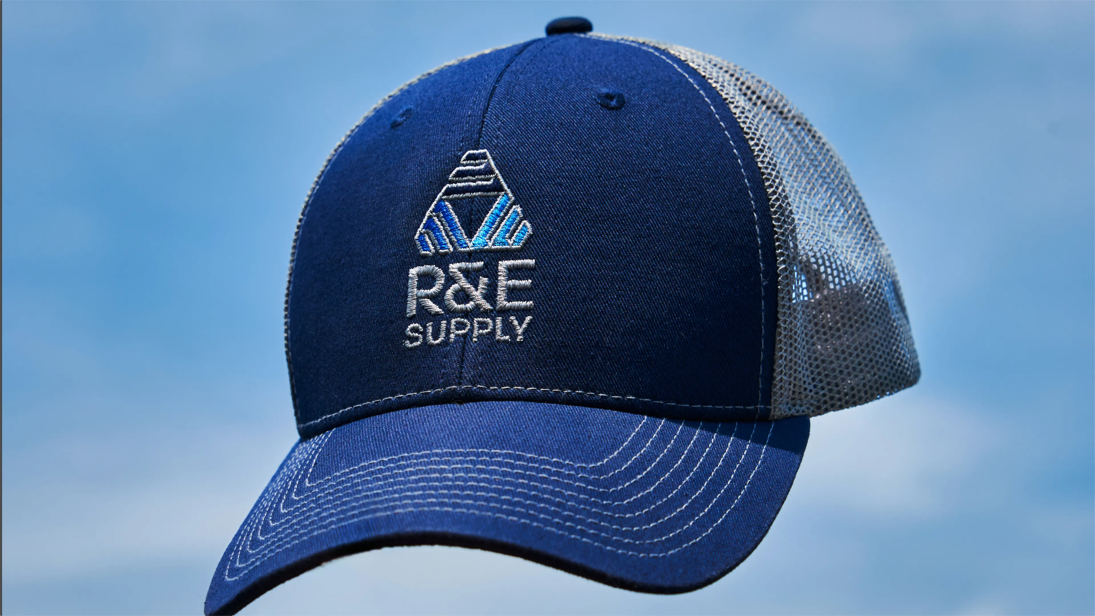 R&E Supply embroidered logo trucker hat in navy and grey mesh against blue sky, brand identity by Unbound Collective