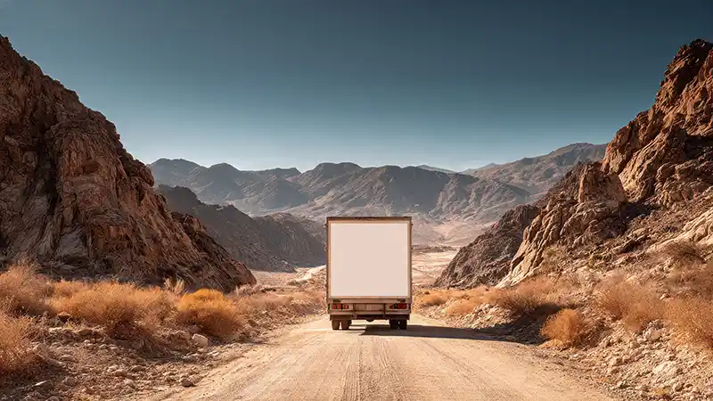 View of back of white box delivery truck driving down a dirt road towards the mountains