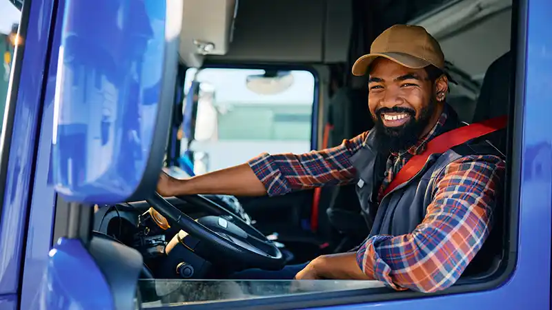 Smiling African American truck driver behind steering wheel in cabin looking at camera.