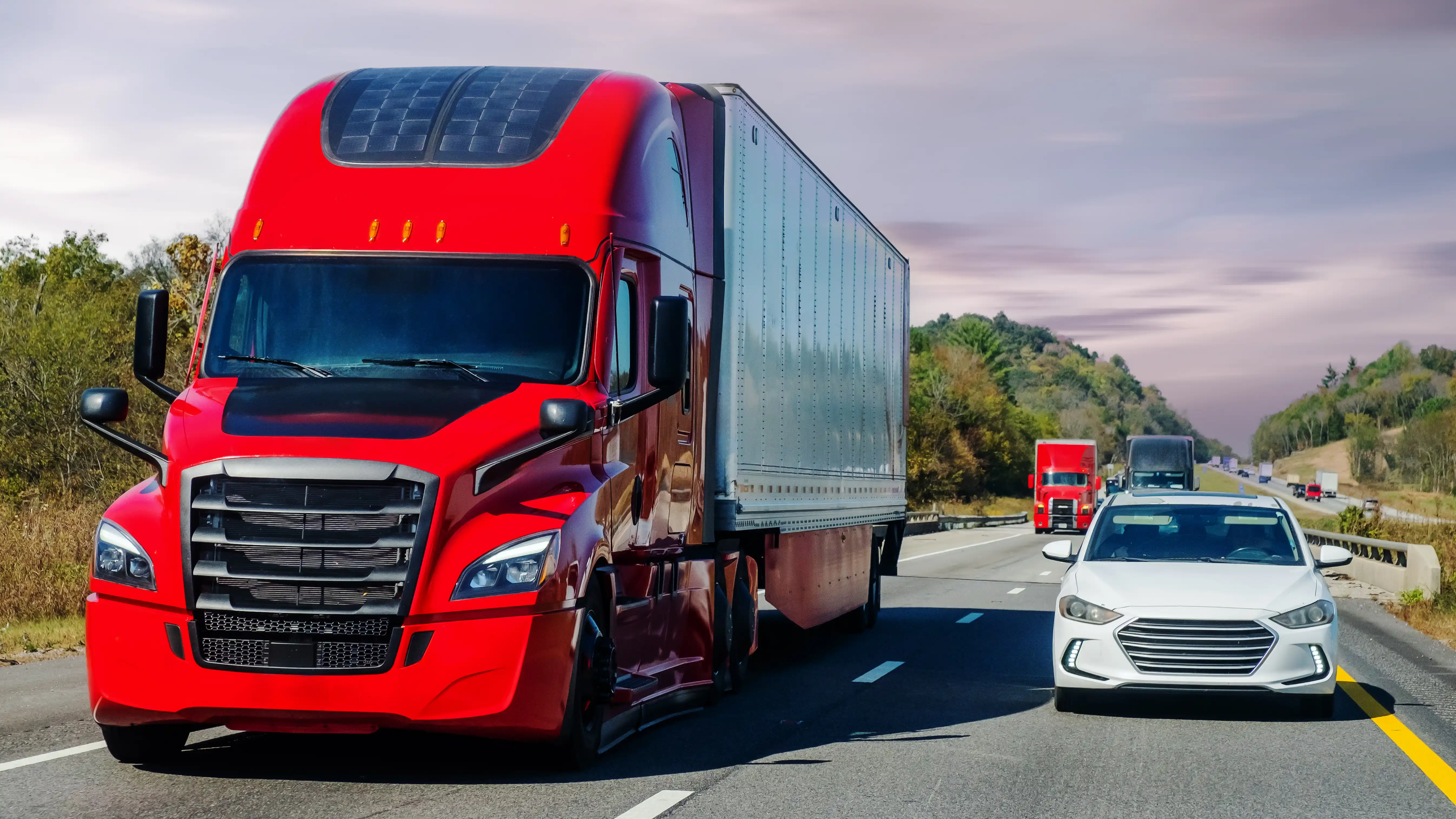 Red semi truck driving down the road with white passenger car driving ext to it.