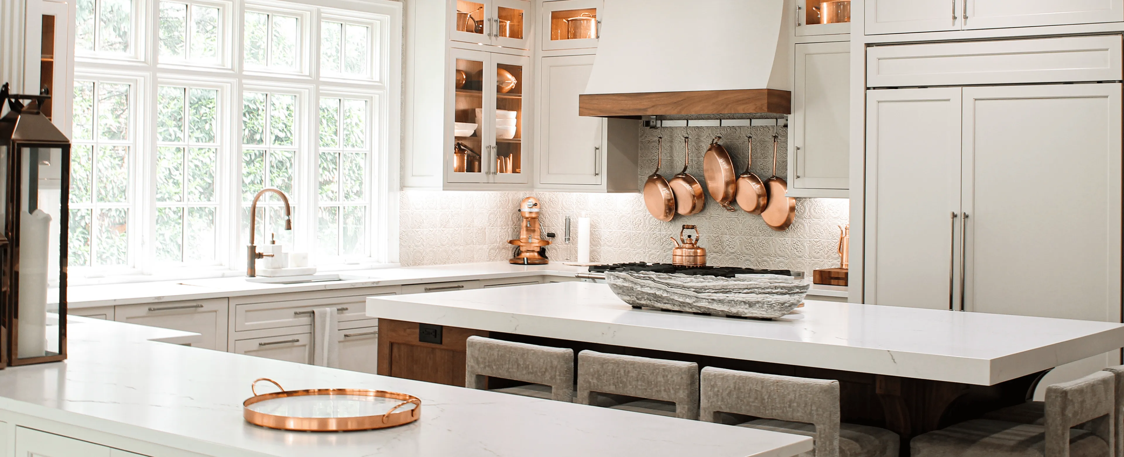 Modern kitchen with white cabinetry, marble countertops, copper cookware hanging above the stove, and a large window letting in natural light.