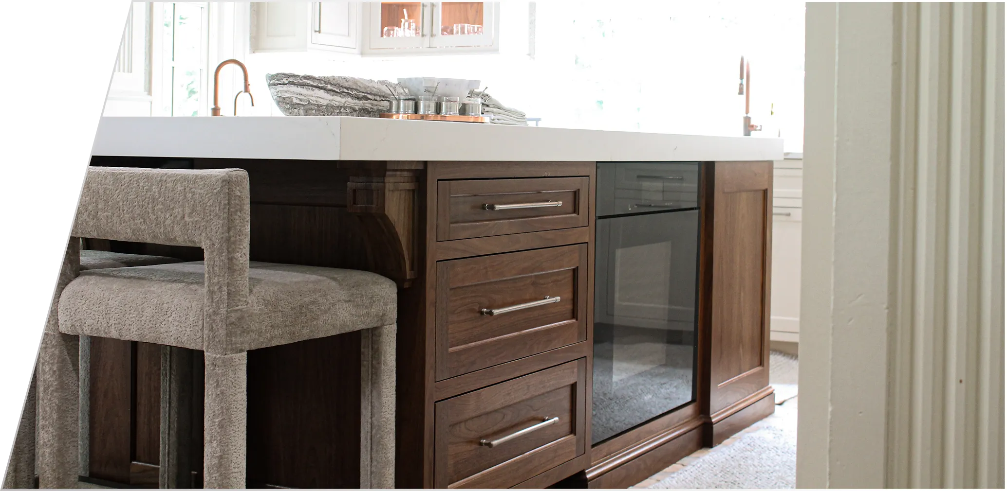 A kitchen island with wooden drawers, a built-in glass-front cabinet, white countertop, and a gray upholstered chair.