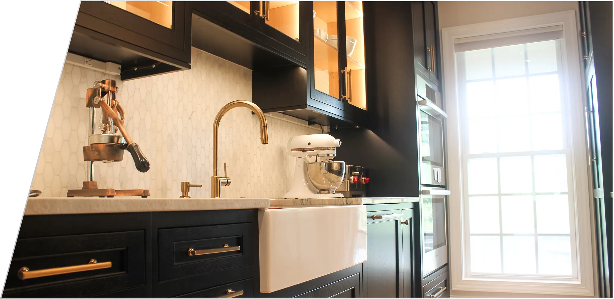 Modern kitchen with black cabinets, a white farmhouse sink, gold faucet, espresso machine, stand mixer, and large window letting in natural light.