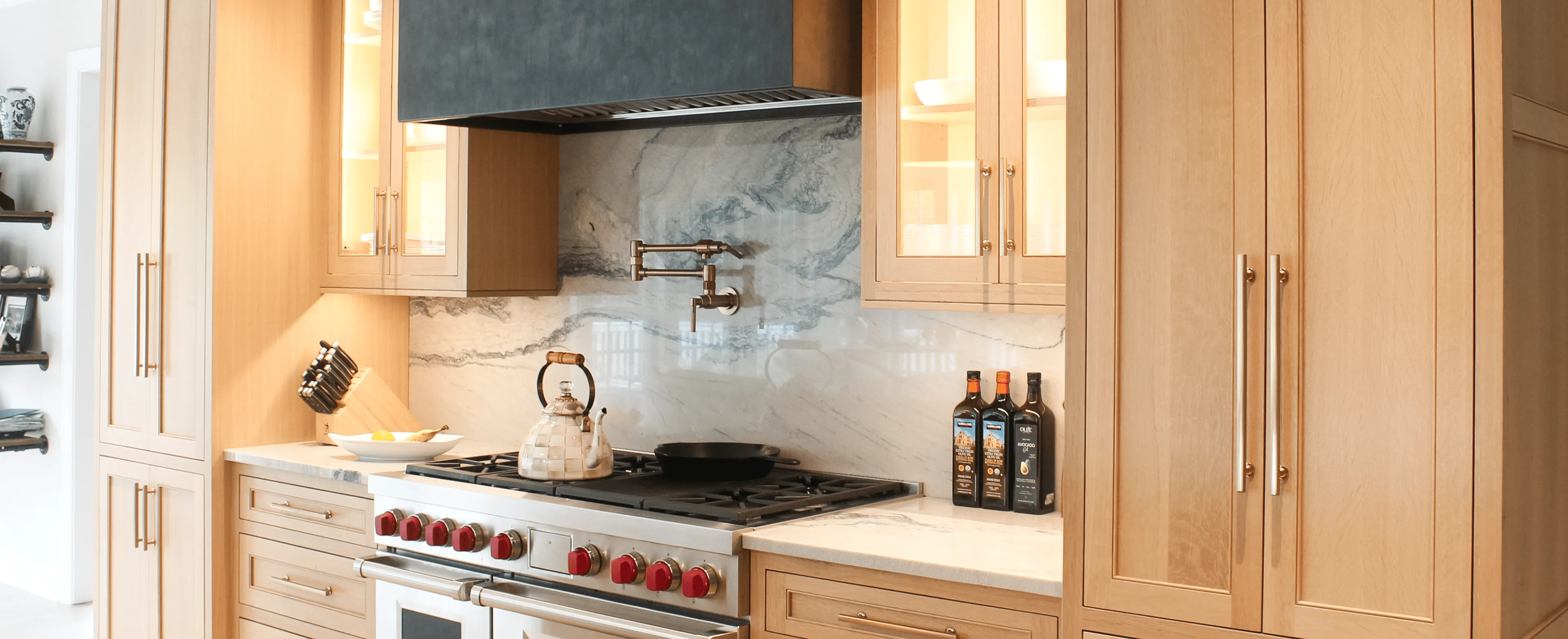 Modern kitchen with light wood cabinets, marble backsplash, stainless steel stove, pot filler, kettle, and bottles of olive oil on the counter.