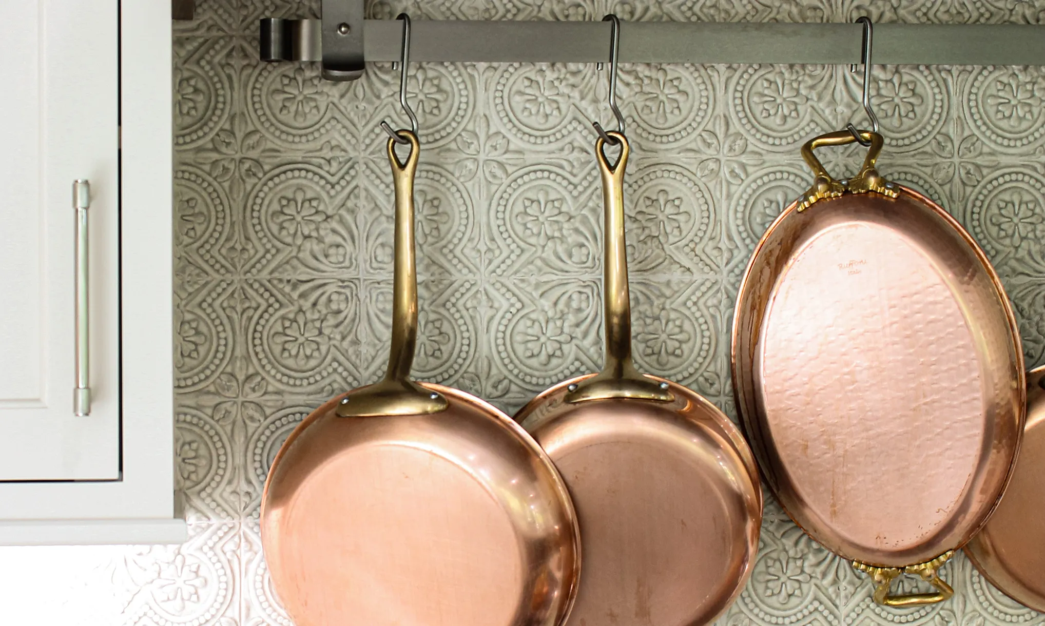 Three copper pans with brass handles hang from hooks on a wall-mounted rack against a decorative patterned backsplash.