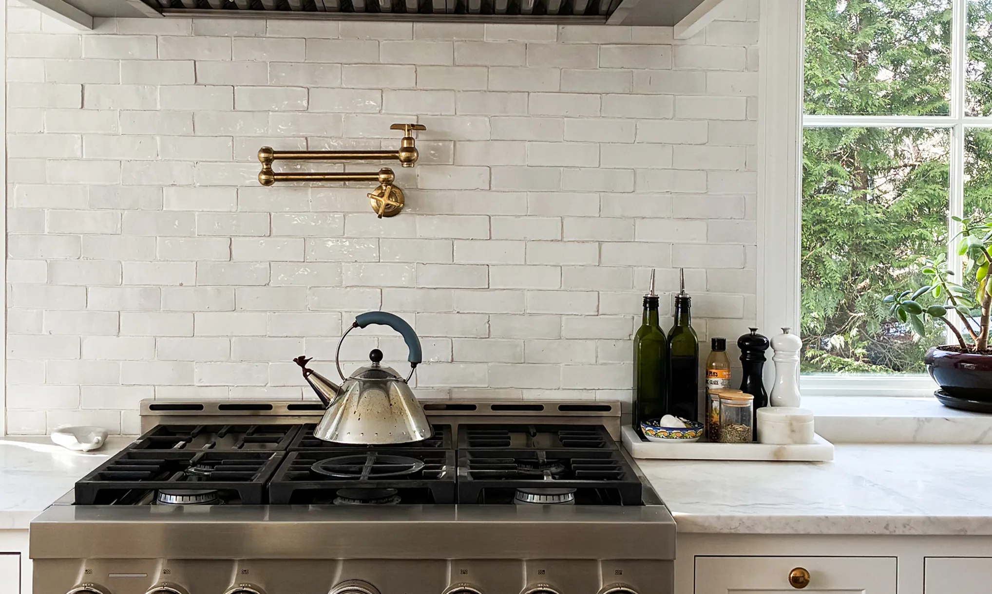 A stainless steel stove with a kettle on a burner, beneath a pot filler faucet on a white tile backsplash.