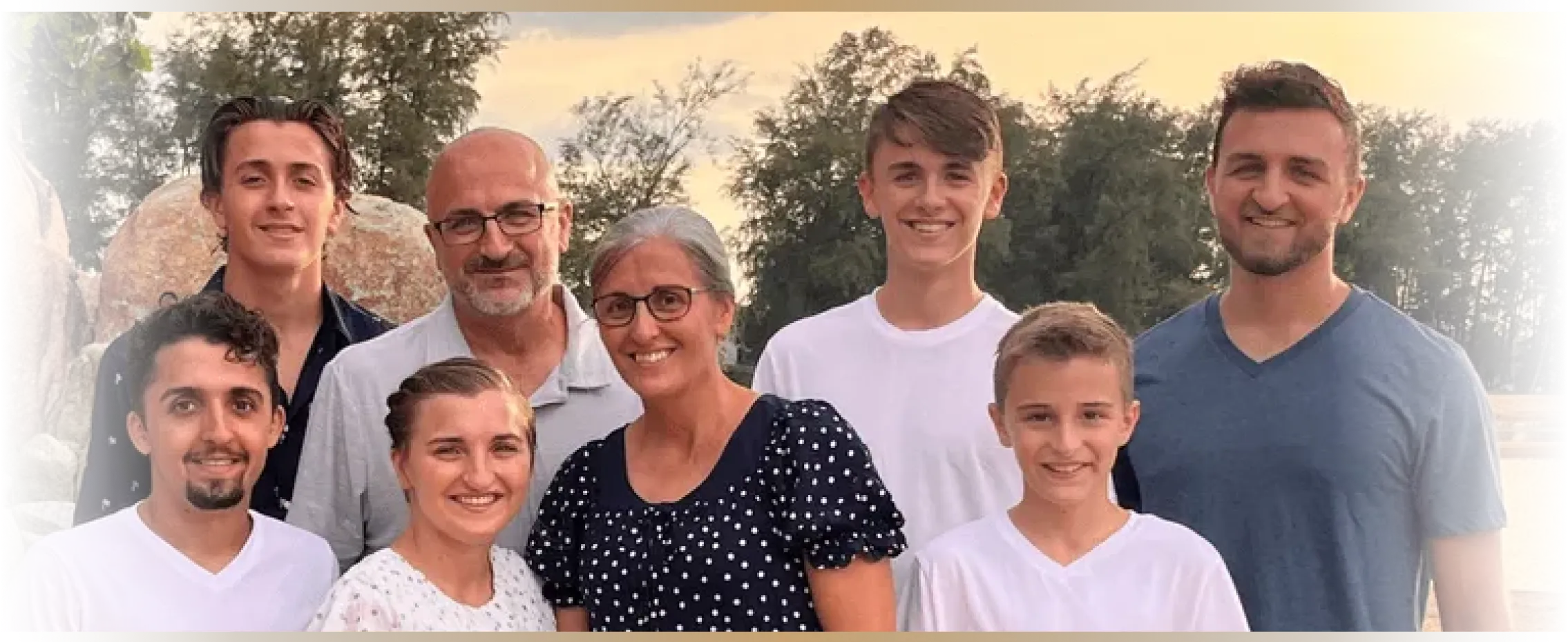The Twin Valley family including four adults and four children or teens, pose outdoors in front of trees at sunset.