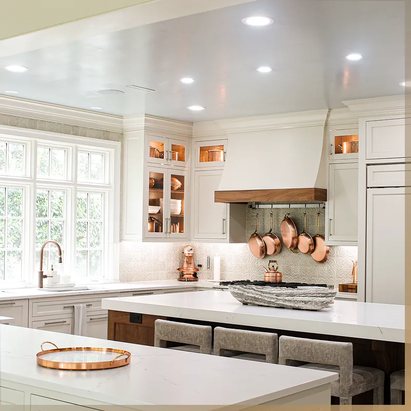 Modern kitchen with white cabinets, marble countertops, a large island with stools, copper pots hanging above the stove, and large windows providing natural light.
