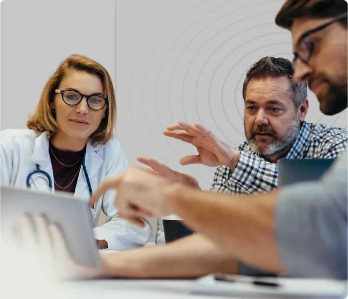 Three professionals engaged in discussion with a laptop, one woman wearing a doctor's coat and stethoscope.