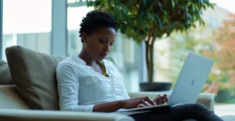 Woman sitting on a couch typing on a laptop near large window with a green plant behind her.
