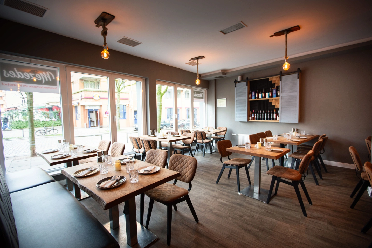 Heller Gastraum des griechischen Restaurants Mezedes in Hamburg mit eingedeckten Tischen, großen Fensterfronten und Weinregal.