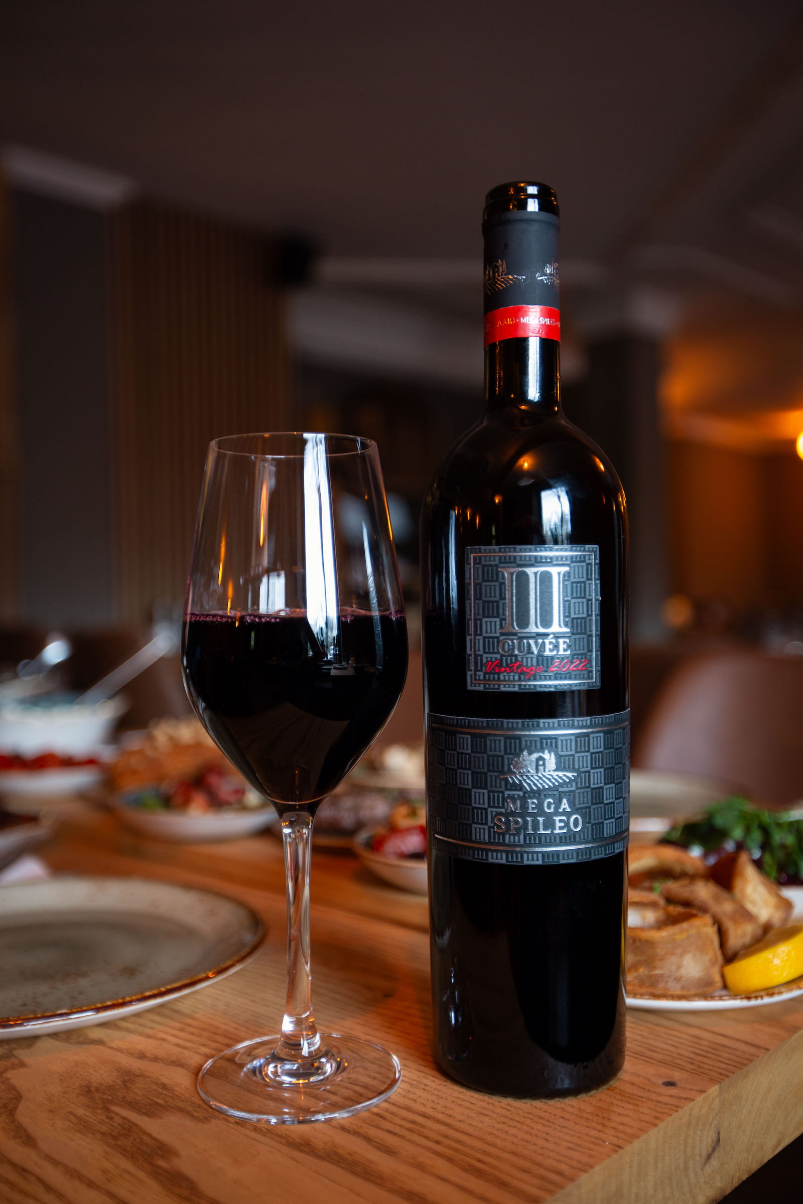 Flasche Mega Spileo Cuvée Rotwein mit gefülltem Weinglas auf einem gedeckten Tisch im griechischen Restaurant Mezedes.