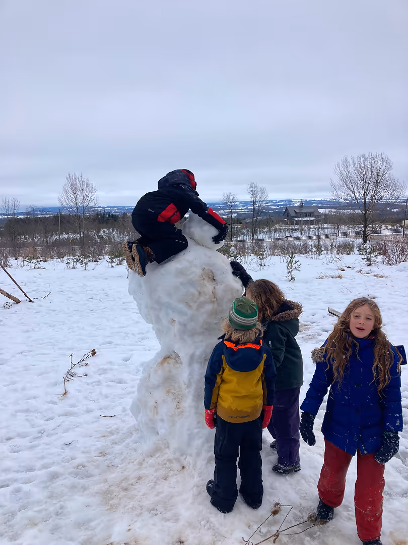 Four children outdoors in winter around a large melting snowman, with one child climbing on top and the others standing nearby on snowy ground.