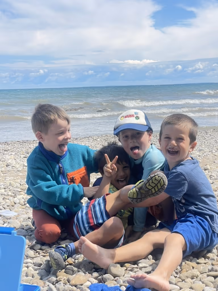 Four boys sitting and laughing together on a rocky beach with the ocean and cloudy sky in the background.