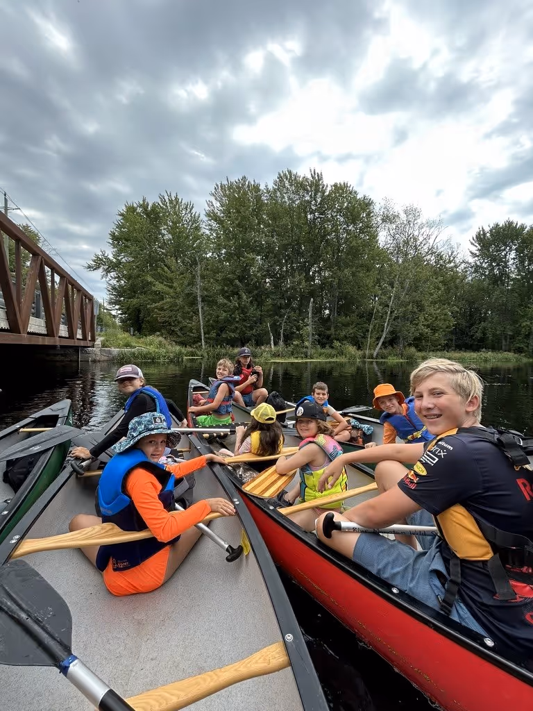 Group of children wearing life jackets sitting in canoes on a calm river near a wooden bridge and trees under a cloudy sky.