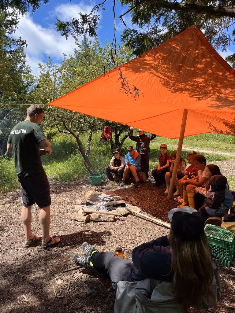 Group of children sitting under an orange tarp in a forest, listening to an instructor near a campfire.