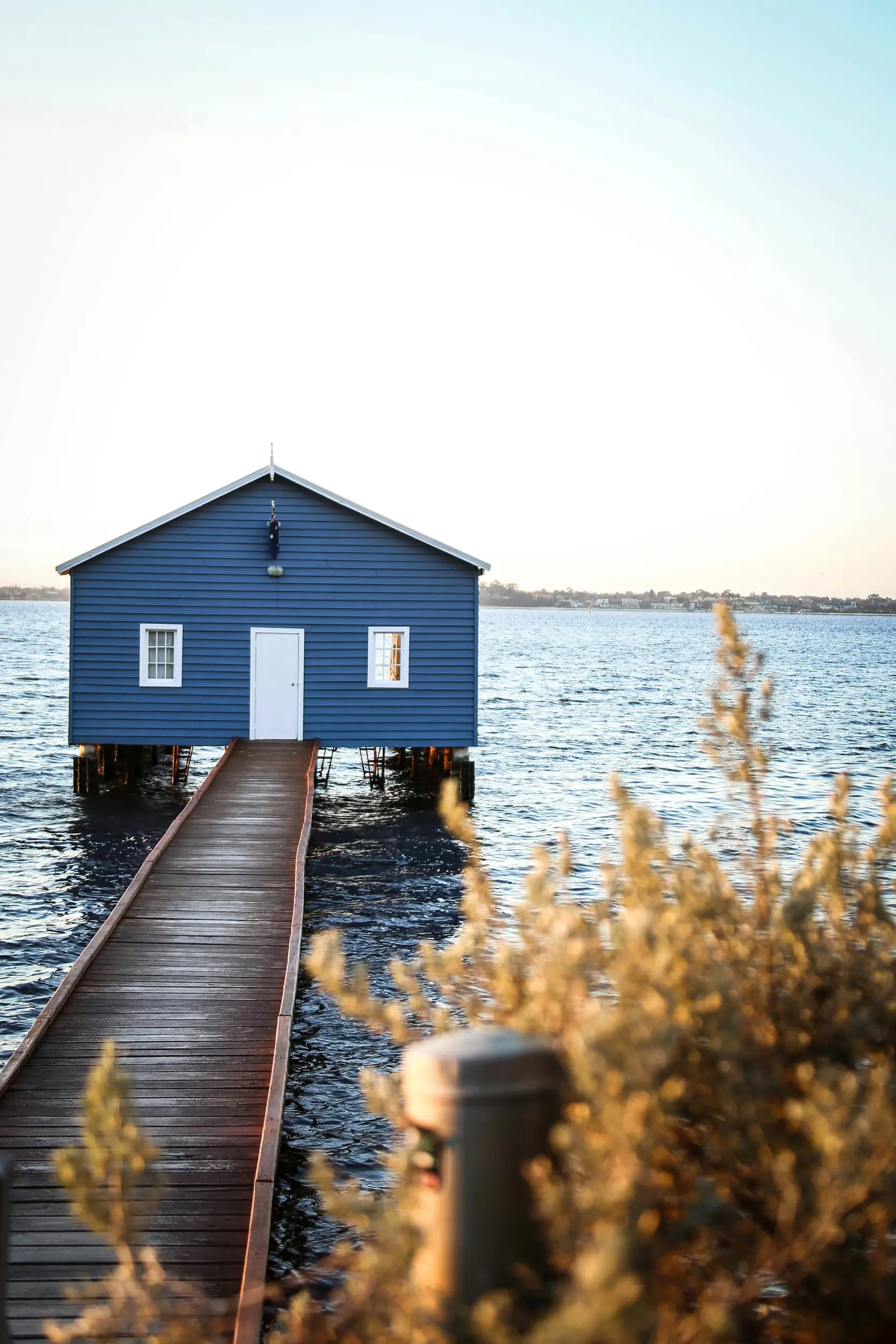 Blue boathouse on water connected by a wooden walkway with dried plants in the foreground.