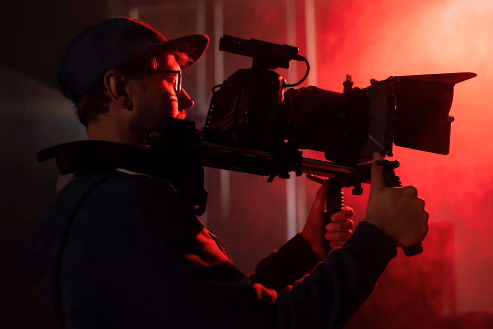 Kameramann bedient professionelle Filmkamera auf Stativ bei Nacht mit bunten Lichtern im Hintergrund.