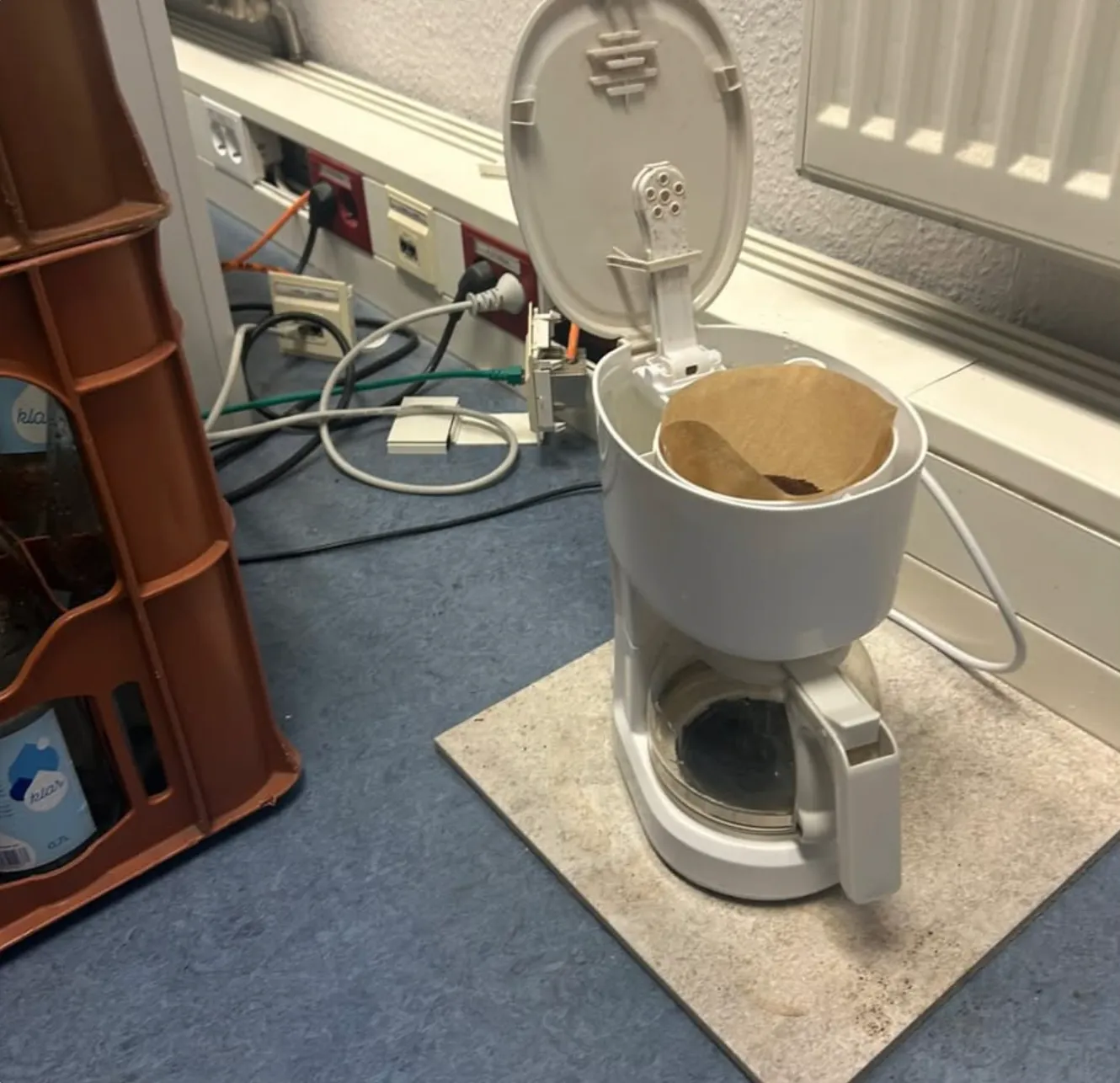 Weiße Kaffeemaschine mit offenem Deckel und braunem Papierfilter, steht auf einer hellen Unterlage neben einer Steckdosenleiste mit mehreren Kabeln.