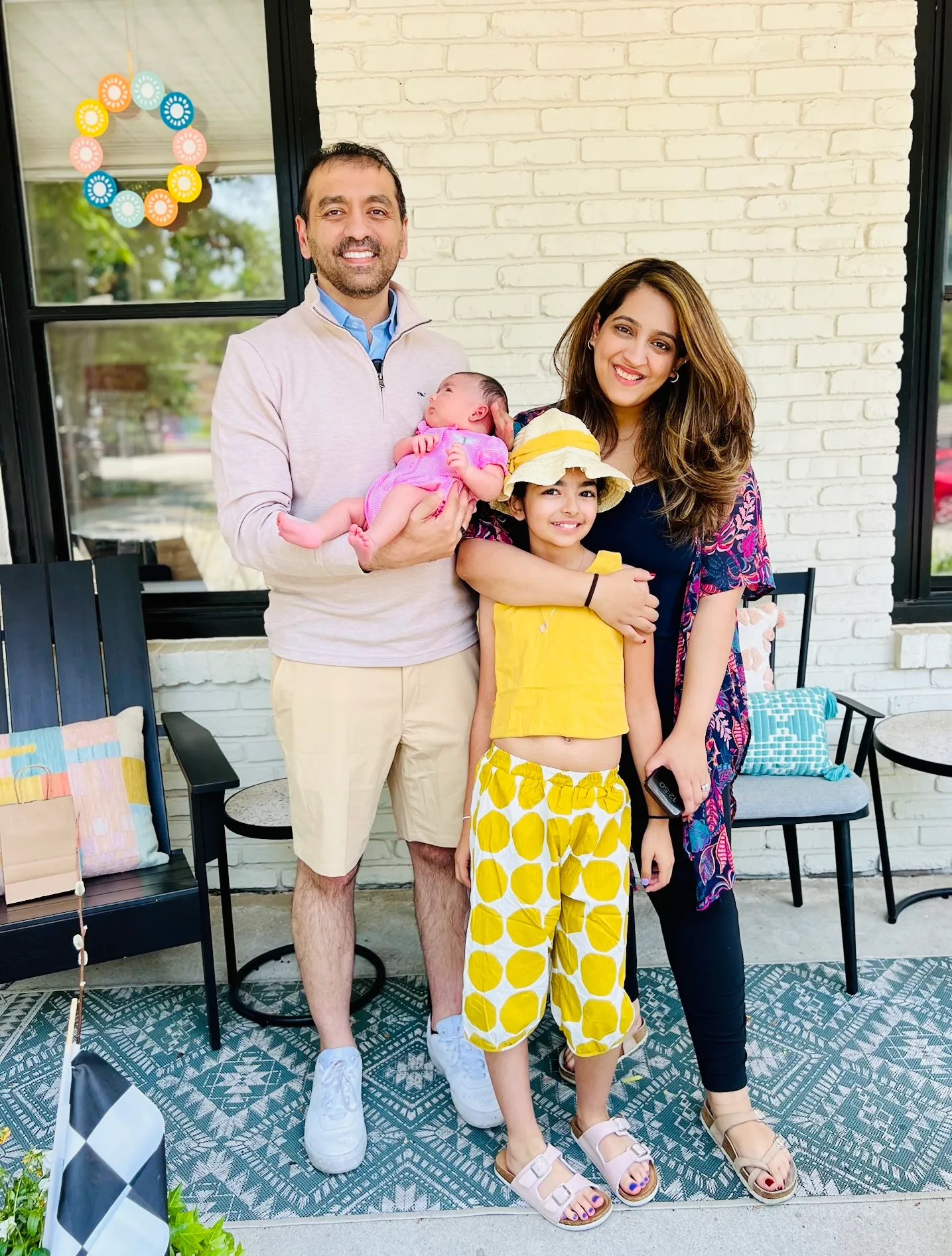 Smiling family of four standing on a porch with a man holding a baby, a woman and a girl in yellow outfit and hat.