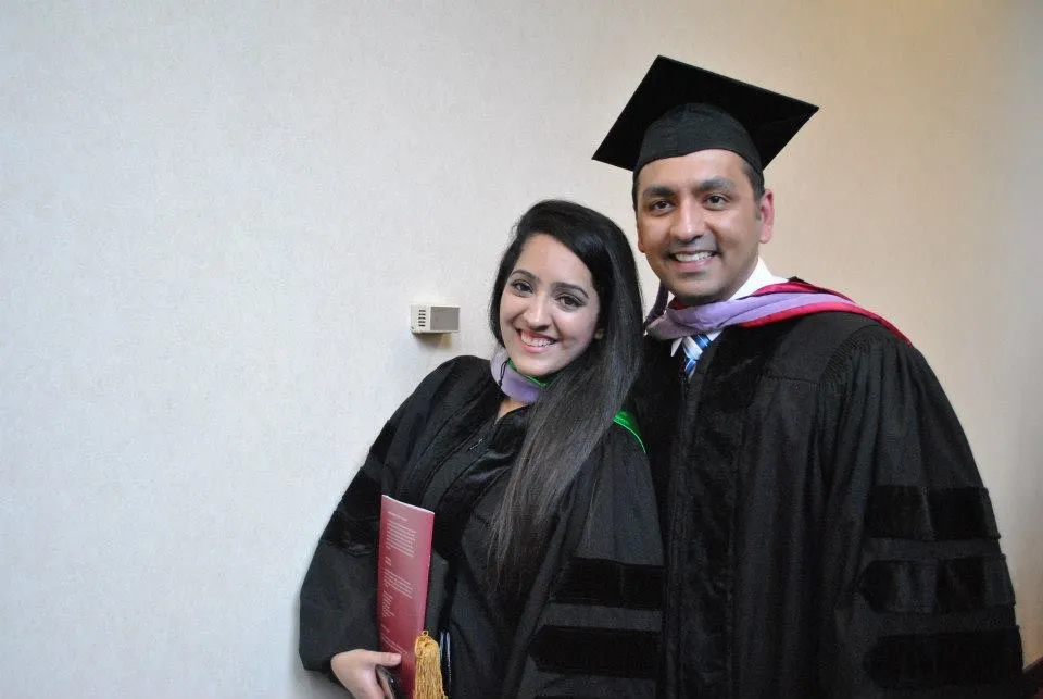 Two smiling graduates in black academic gowns and hoods posing together against a plain light wall.